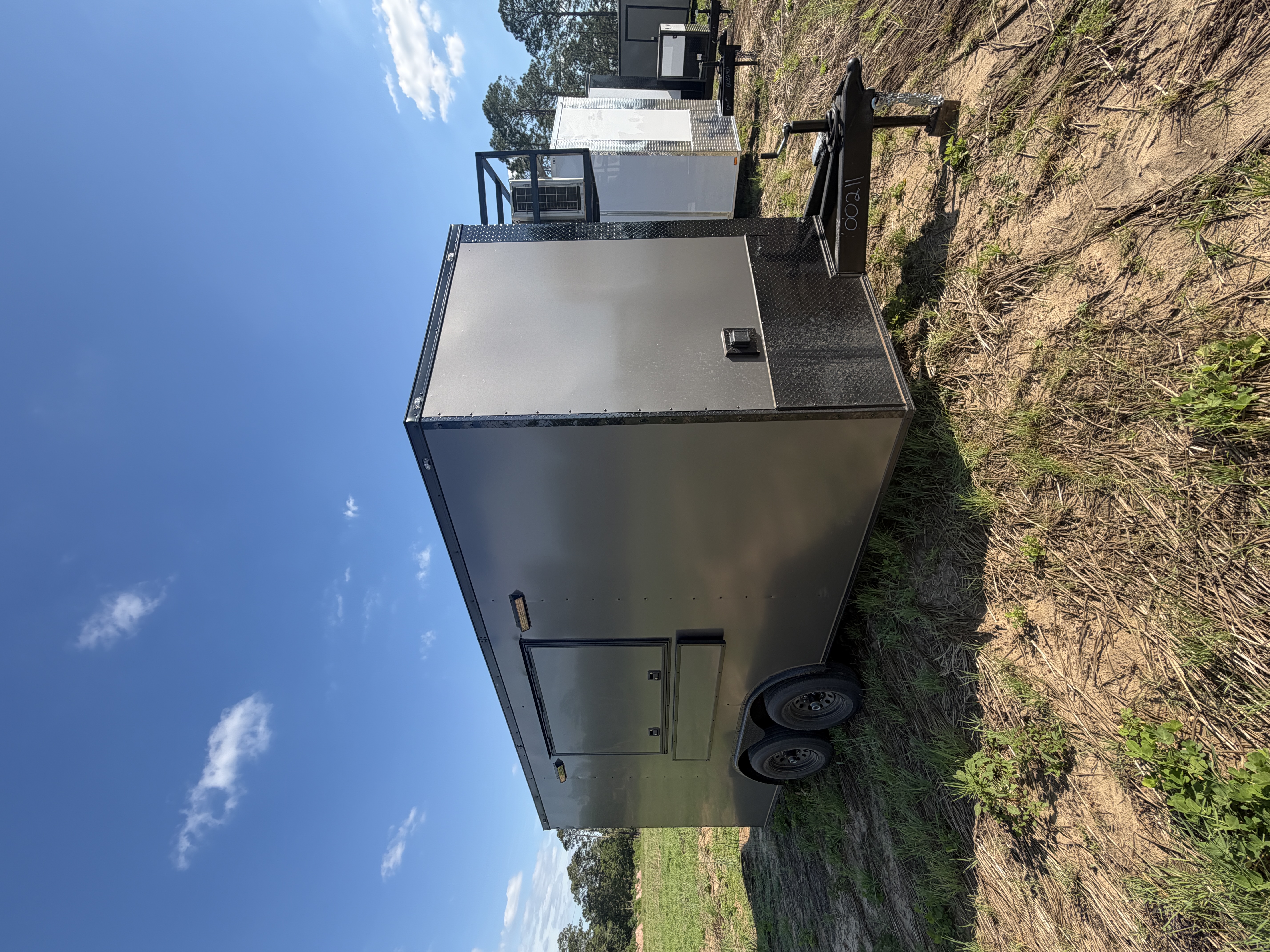 Charcoal grey 8.5x16 tandem-axle concession trailer with full blackout trim, diamond-plate stone guard, side concession service window, and extended tongue — parked in a grassy field