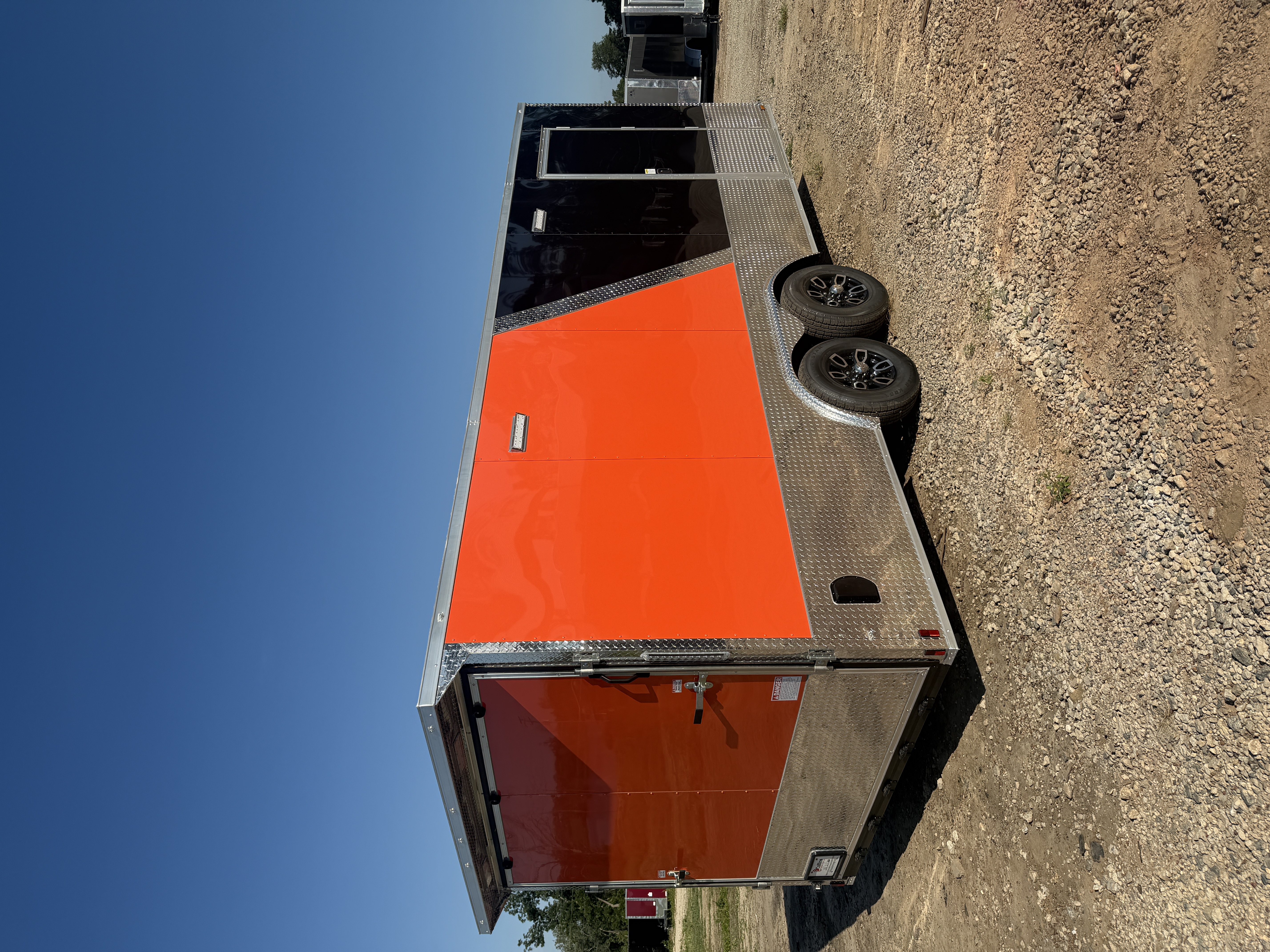 Two-tone orange and black enclosed 8.5x20 tandem axle car hauler — driver-side rear three-quarter view showing the diagonal split between the glossy orange front and black rear, silver ATP diamond-plate trim wrapping the base and slanted V-nose, black aluminum mag wheels, and a rear-corner man door, parked on a gravel lot under a clear blue sky