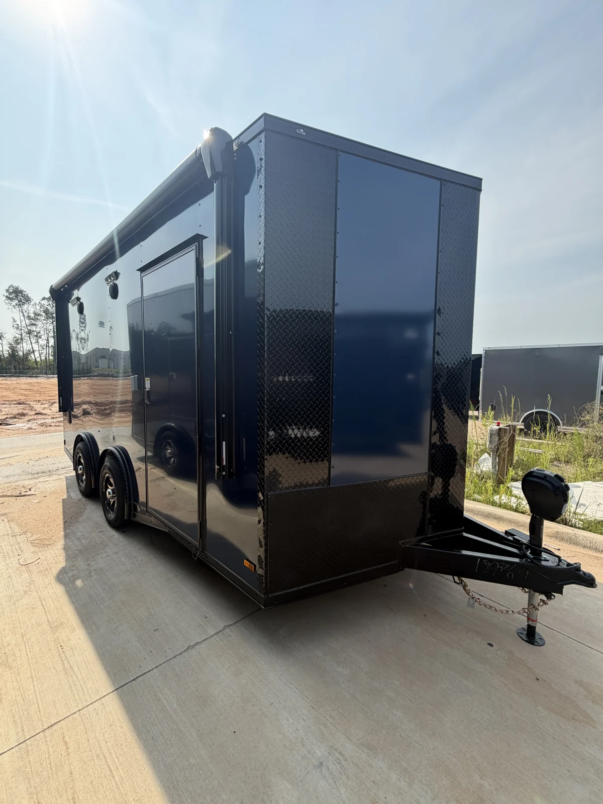 Indigo Blue 8.5x16 tandem-axle motorcycle race trailer viewed from the front corner on a concrete lot, showing black trim, a rolled-up awning, side entry door, spread axles, and electric tongue jack