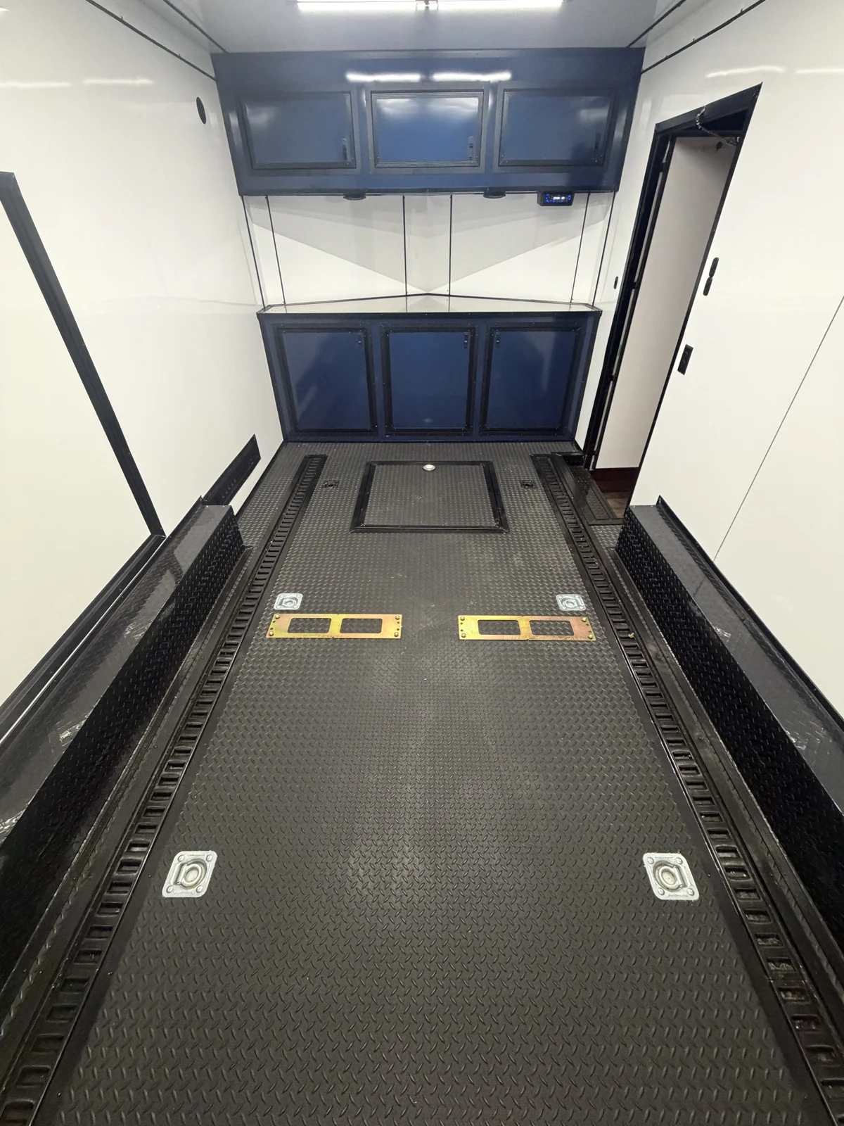 Motorcycle trailer interior showing floor-mounted motorcycle backer plates, recessed floor E-track, a tire compartment lid, and Indigo Blue base and overhead cabinets