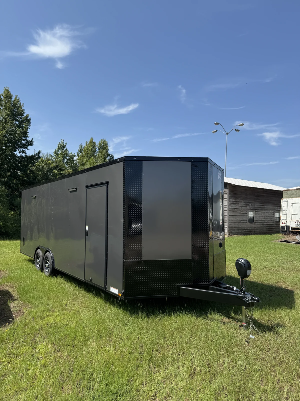 Charcoal Gray 8.5x24 tandem-axle Blackout race trailer viewed from the front corner on grass, showing a black diamond-plate nose, side entry door, Star Mag wheels, extended tongue, and electric jack