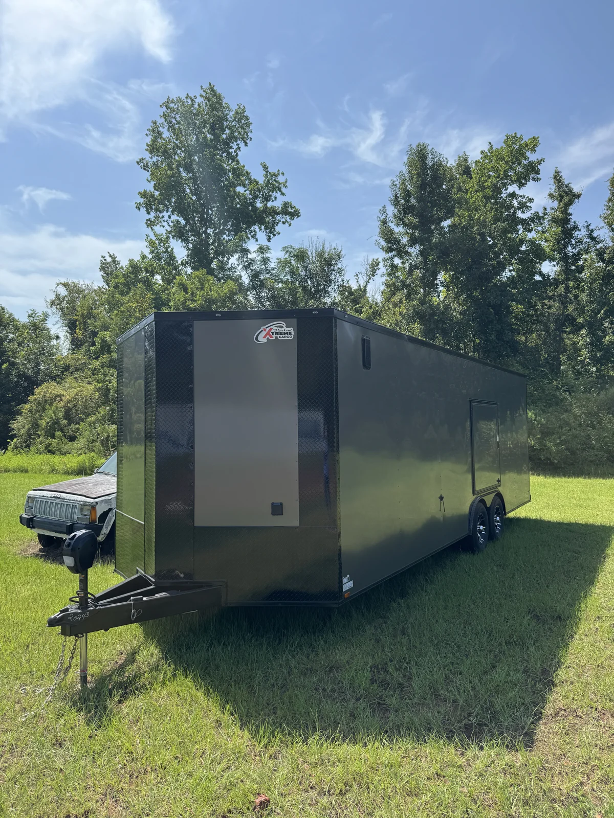 Opposite front corner of the Charcoal Gray Blackout race trailer on grass, showing the electric jack, extended tongue, escape door, and tandem wheels