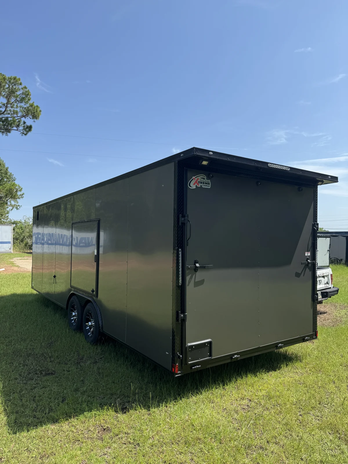 Opposite rear corner of the Charcoal Gray race trailer showing the escape door above the wheels, rear ramp, and black tail wing