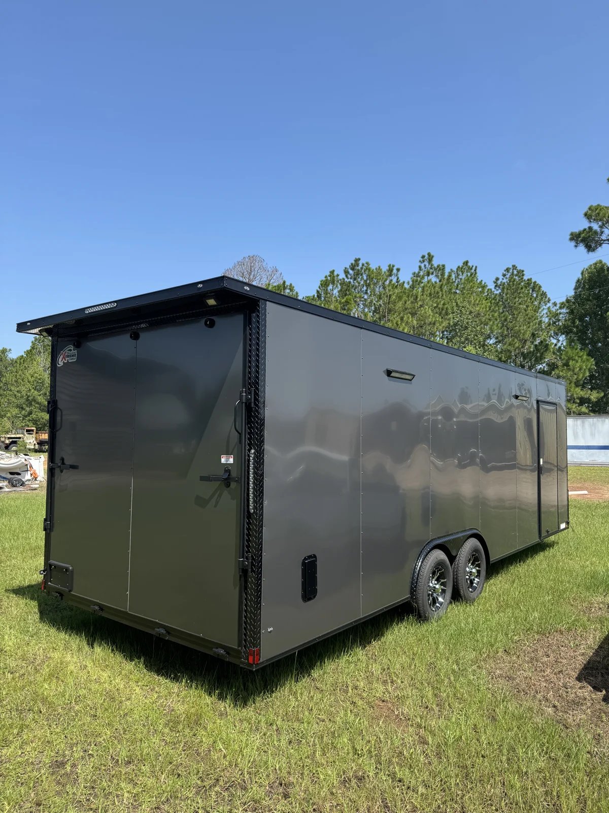 Rear corner of the Charcoal Gray Blackout race trailer on grass, showing the ramp door, tail wing, side entry door, and tandem wheels