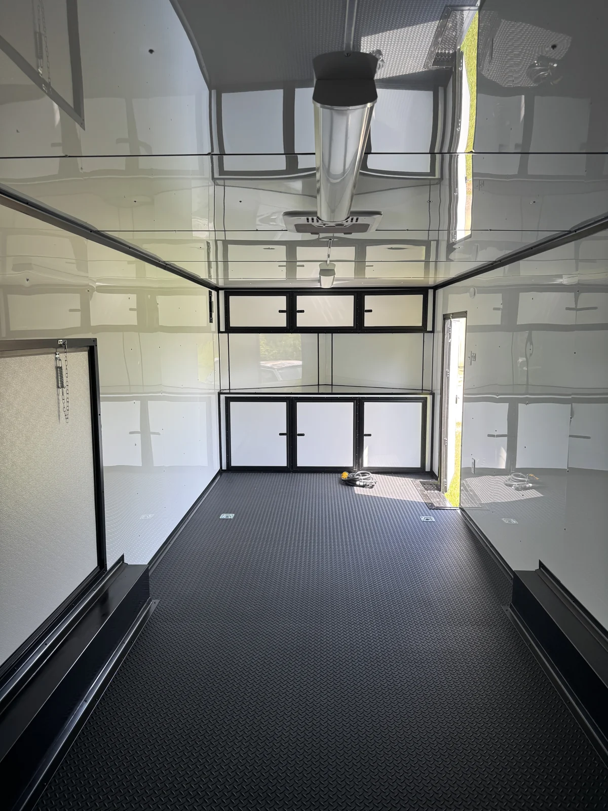 Front-facing race trailer interior showing white cabinets, white walls and ceiling, black trim, rubber tread plate floor, and ceiling-mounted air conditioner