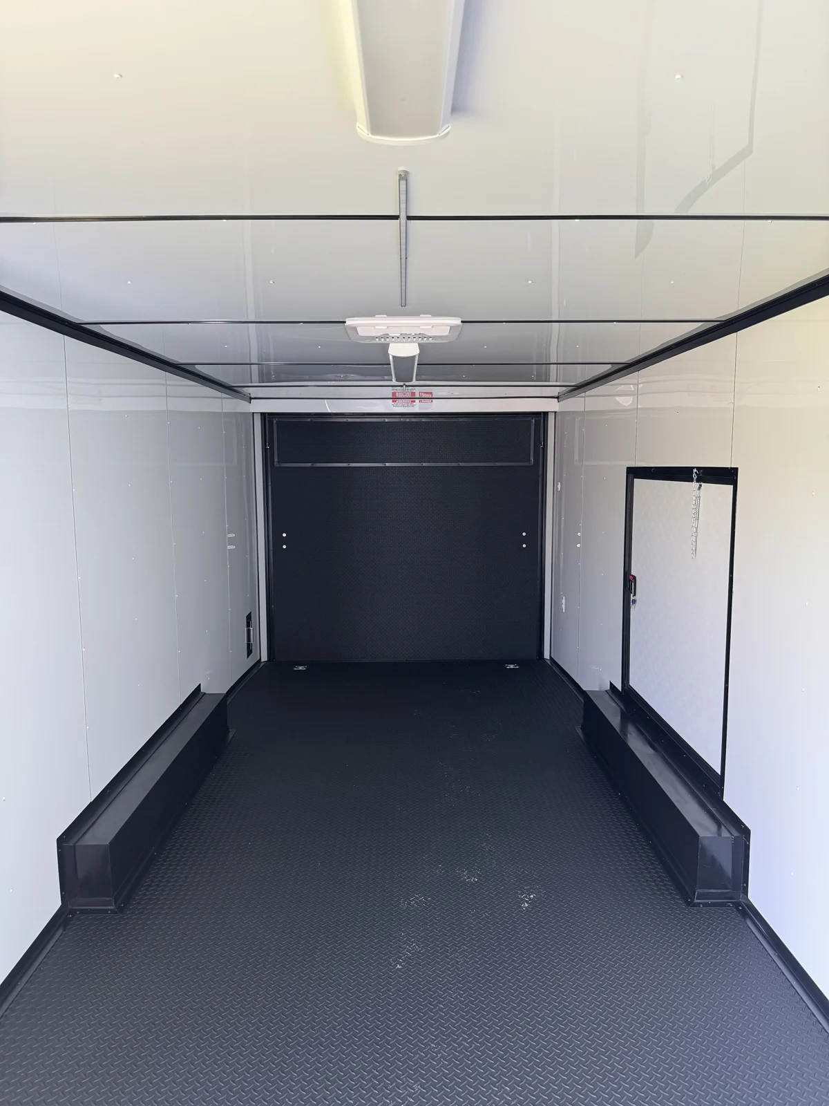 Rear-facing race trailer interior with white walls and ceiling, black trim, rubber tread plate floor, closed ramp, escape door, and ceiling bar lights