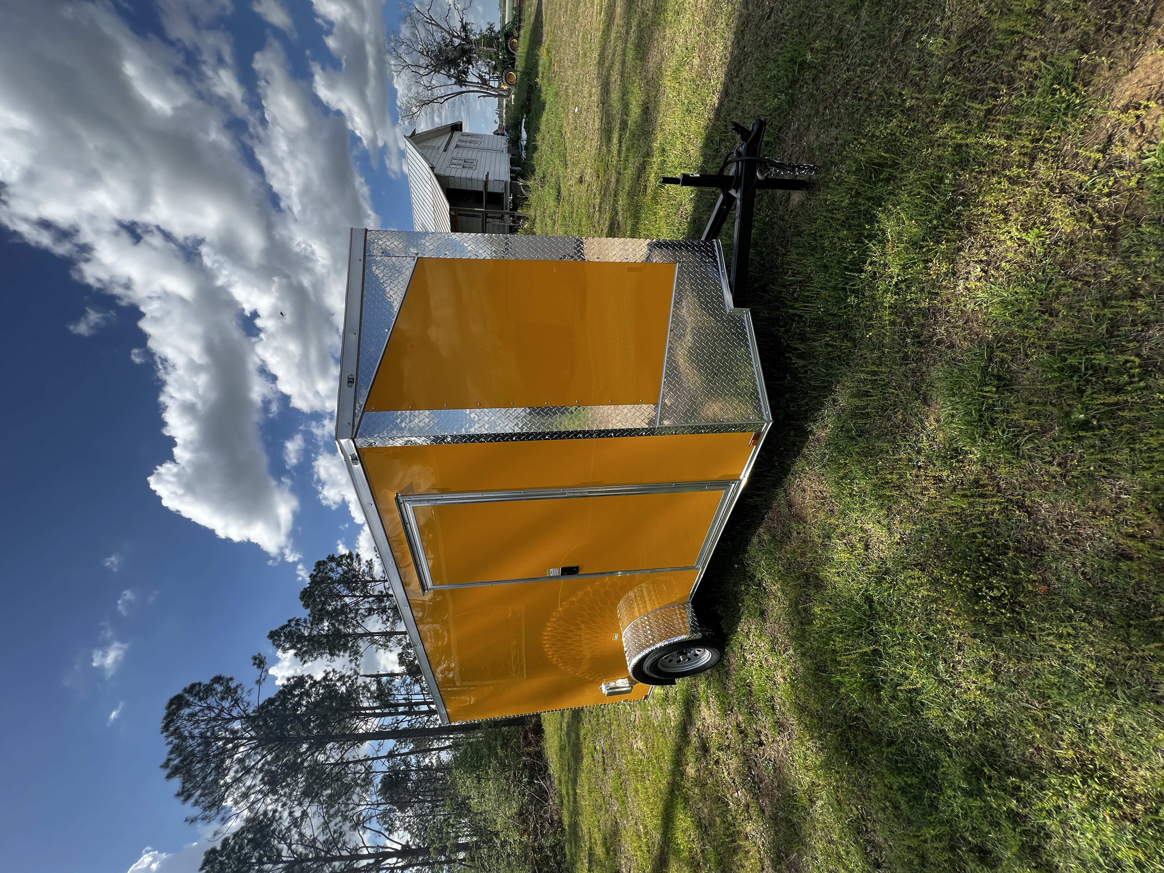 Glossy yellow enclosed 6x12 single axle trailer — driver-side front view showing the slanted V-nose wrapped in chrome ATP diamond-plate trim, chrome corners, silver wheels, and curb-side man door