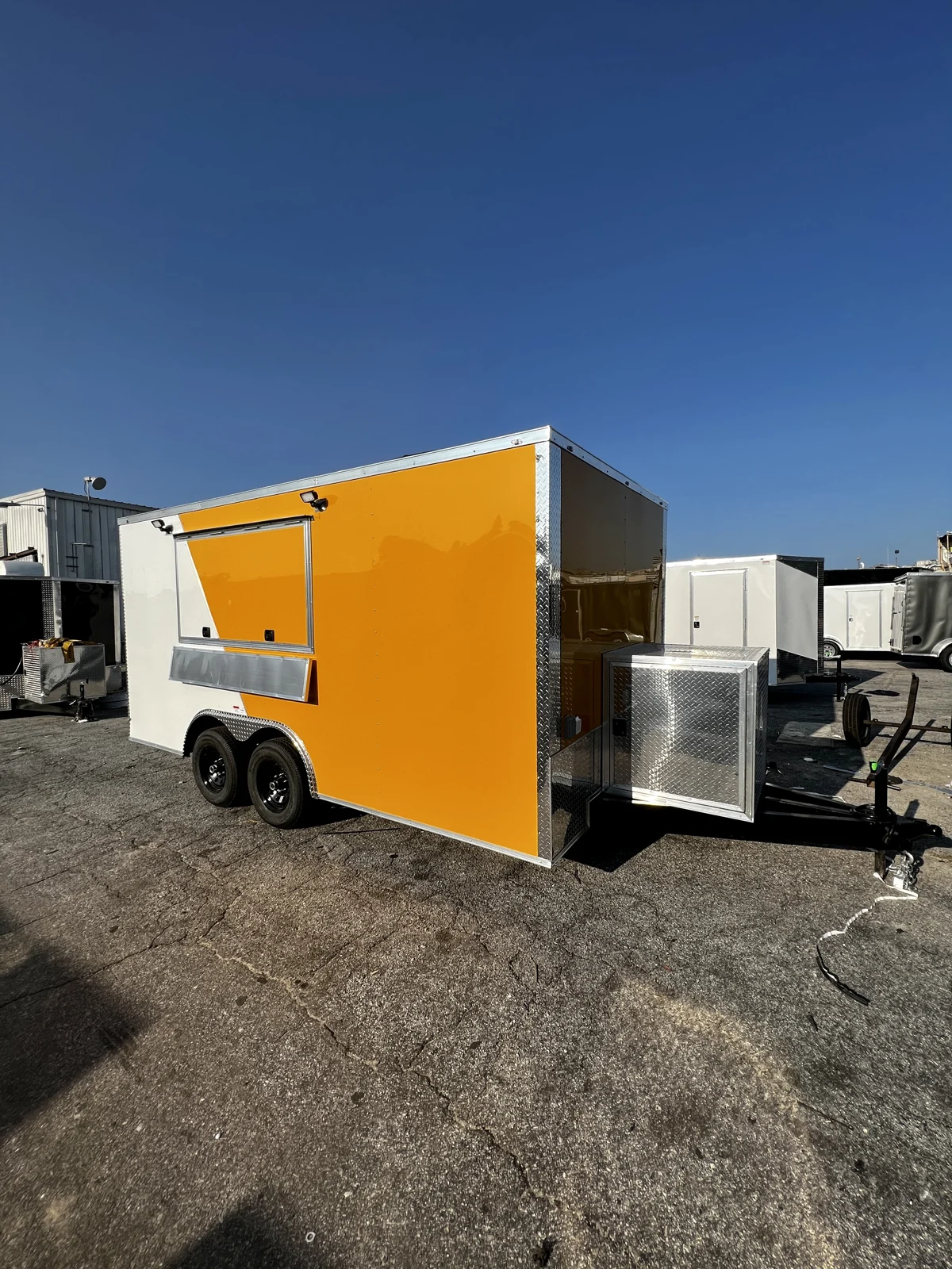 White and yellow split-color 8.5x16 tandem-axle concession trailer viewed from the front corner on a paved lot, showing the concession window, silver trim, black wheels, and diamond-plate generator box on the extended tongue