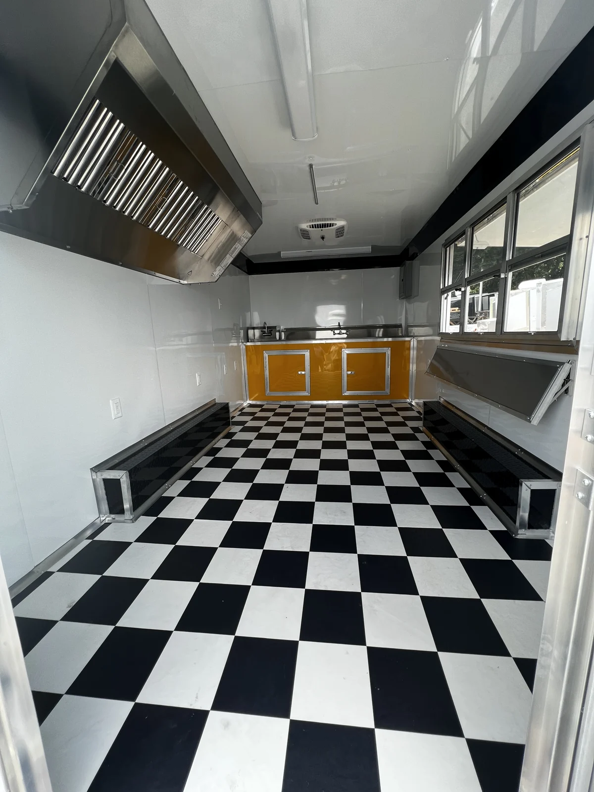 Concession trailer interior with white metal walls and ceiling, checkerboard flooring, hood vent, serving shelf, yellow base cabinets, and ceiling-mounted A/C