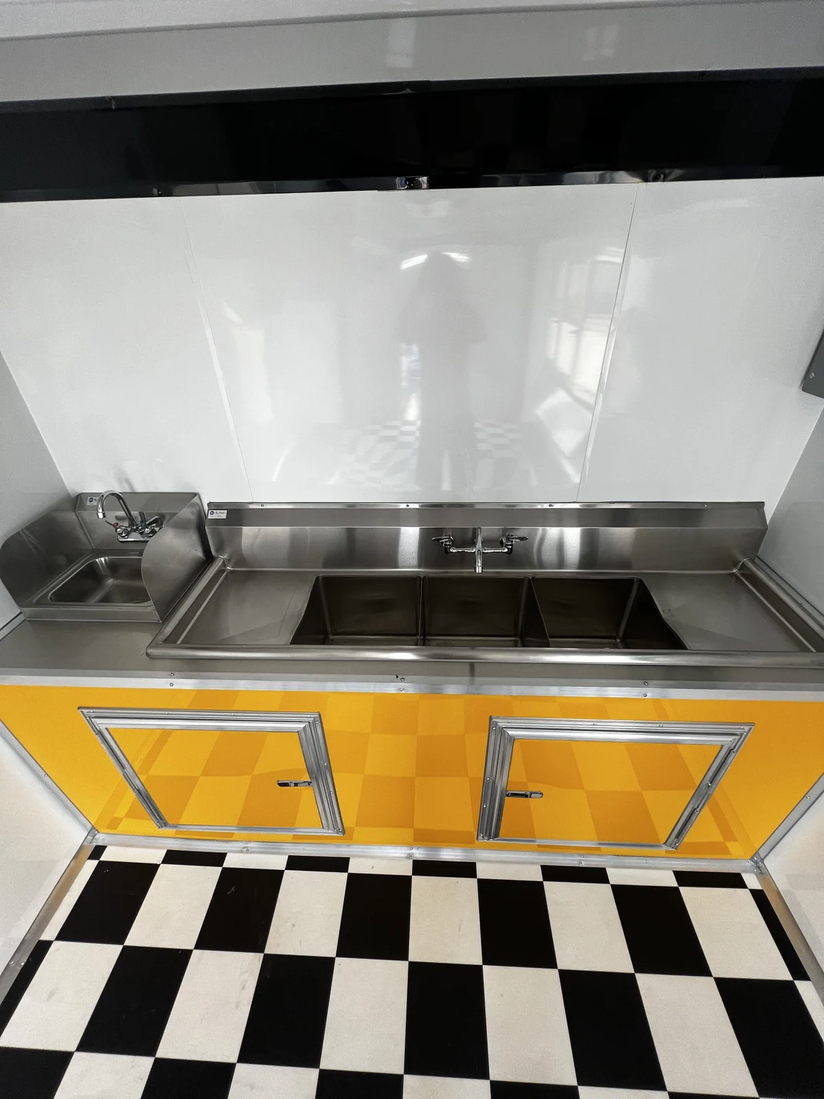Three-bay sink and separate small sink above yellow base cabinets, with white metal walls and checkerboard flooring