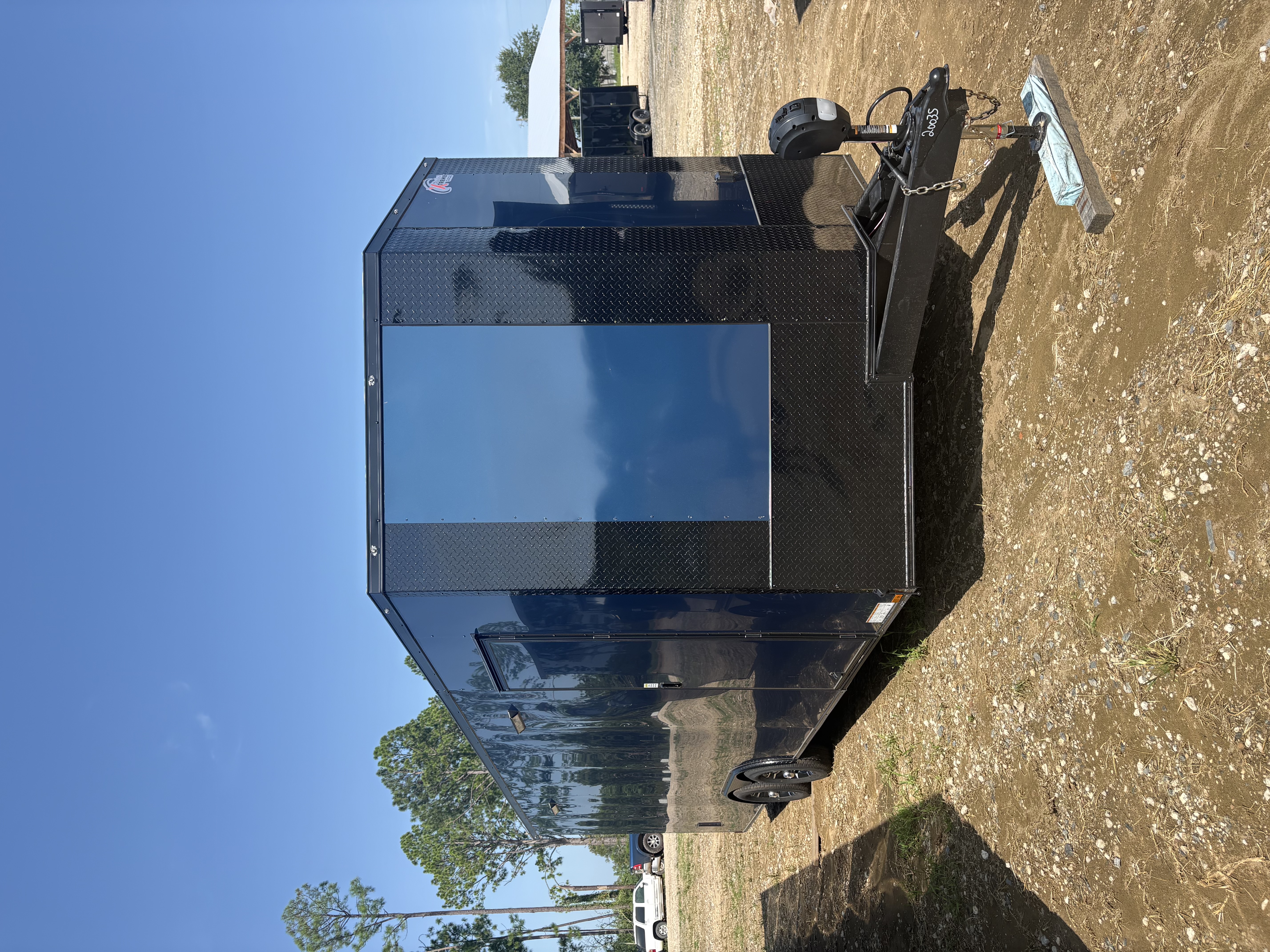 Indigo Blue 8.5x24 tandem axle enclosed trailer on an outdoor dirt lot, front passenger-side view showing blackout diamond-plate nose, extended tongue, electric jack, side door, and Spider Mag wheels