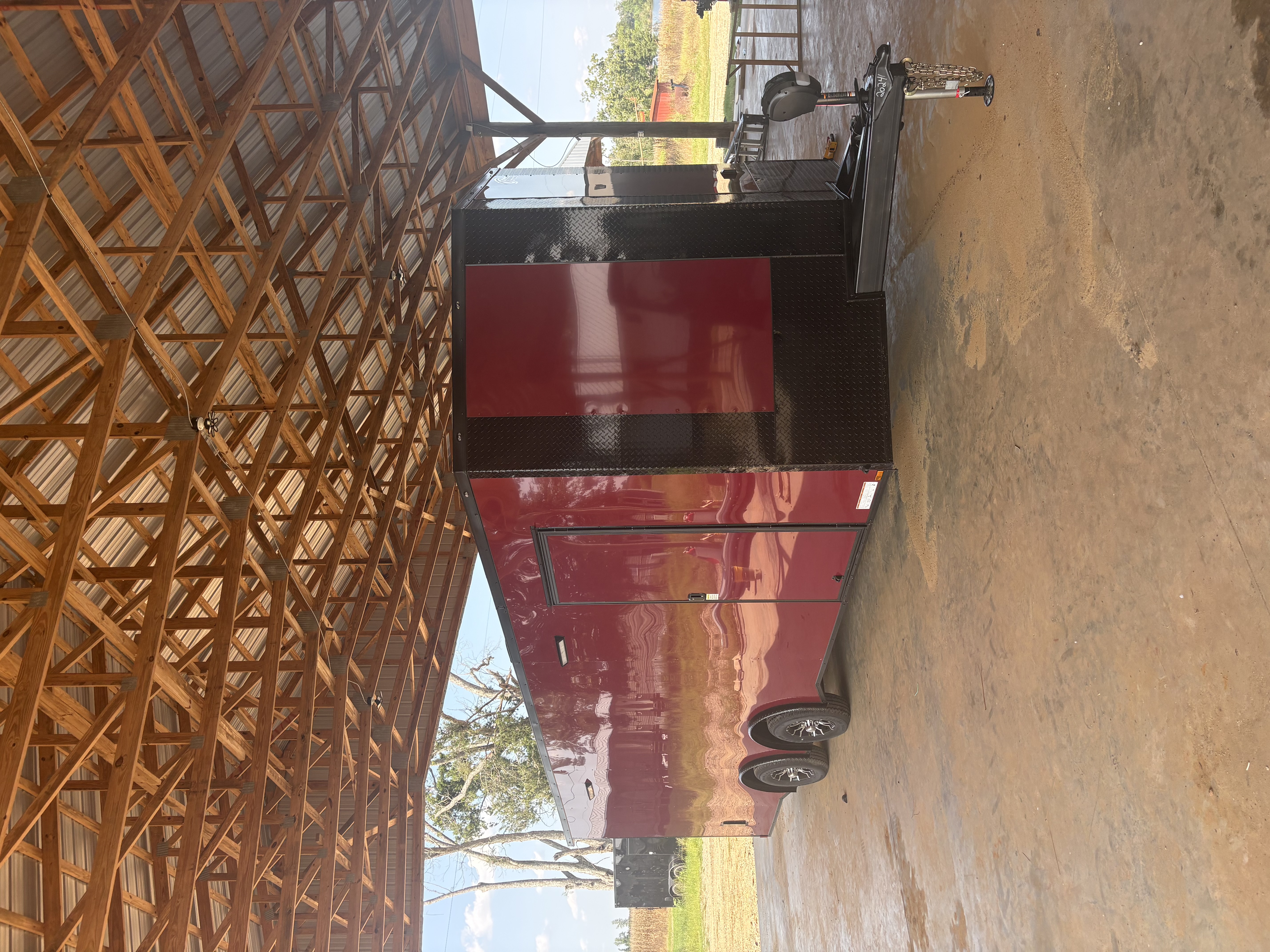 Brandy Wine 8.5x24 tandem axle race trailer beneath a timber-roof shelter, front passenger-side view showing blackout diamond-plate nose, spread axles, Corvette fenders, electric tongue jack, and side door