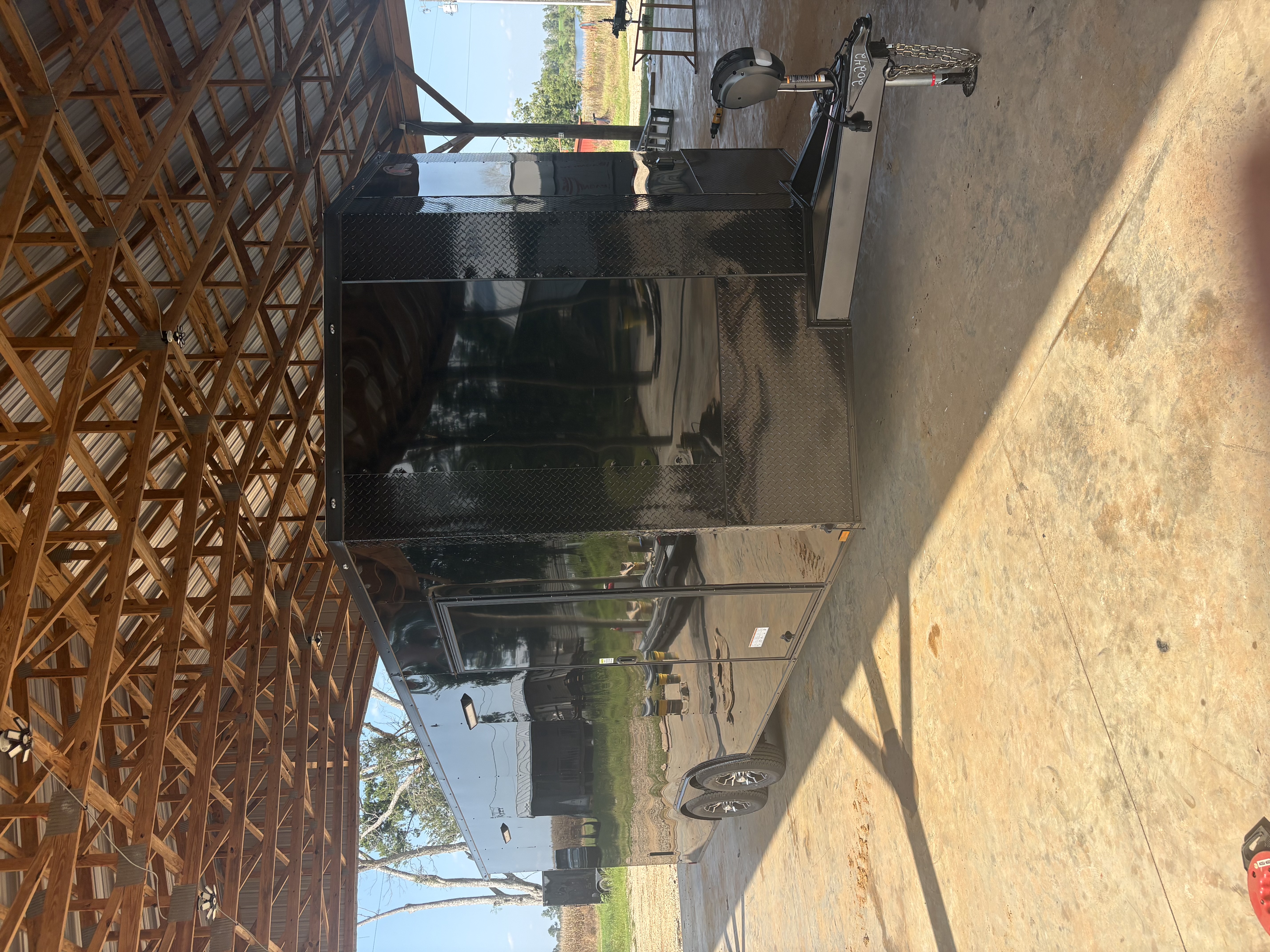 Black 8.5x24 tandem axle enclosed trailer under a timber-roof shelter, front passenger-side view showing blackout diamond-plate nose, extended tongue, electric jack, side door, and Spider Mag wheels